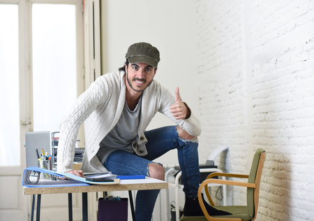 Young trendy businessman in cap and cool hipster look wearing battered denim jeans posing corporate on desk at modern home office in creative freelancer business