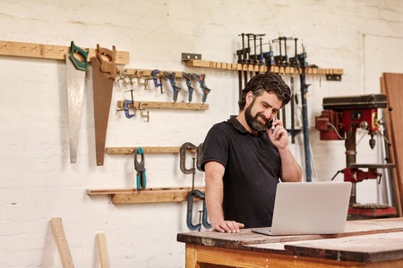 Small business owner standing in his carpentry workshop, smiling while talking on the phone, and looking at his laptop which is resting on his workbench, with tools in rows on the wall behind him