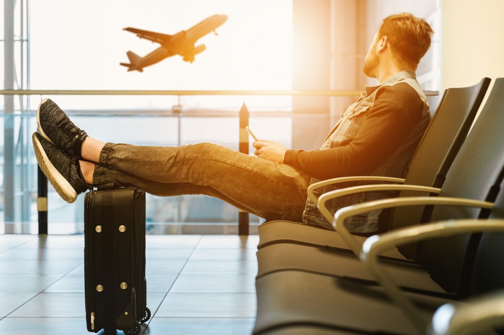 4 tips for less stressful business travel - a young man at airport with his feet up on his luggage watches a jet taking off in the morning sun