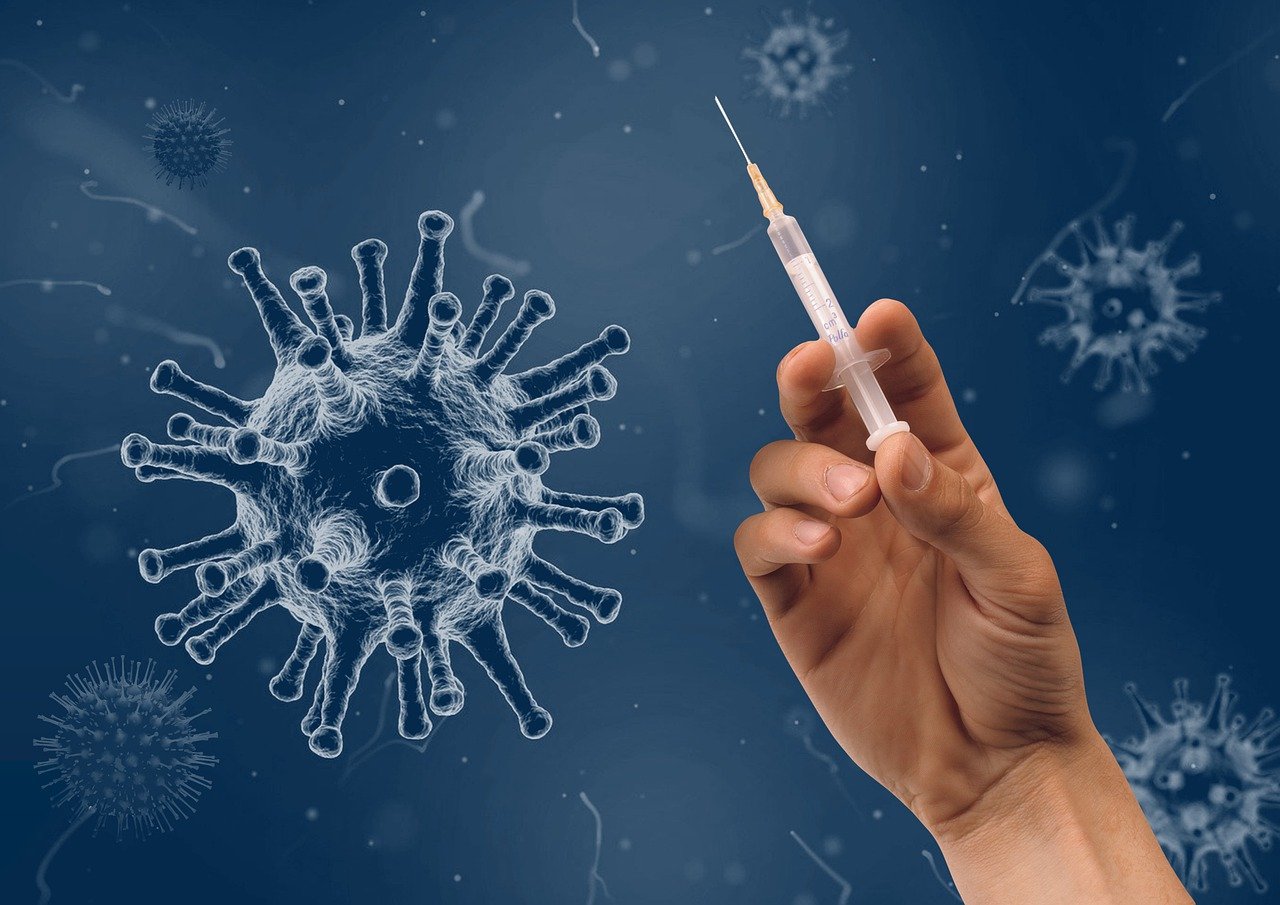 Business Update – 28 February 2022 - a hand holding up a vaccine syringe against a background of COVID-19 viruses.